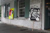 A photograph of the outside of a brick building, with two signs standing against the wall, between the building's front windows, on the porch. The signs depict stylized figures, one saying 