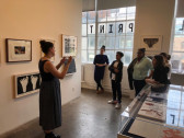 Several students listen to a woman speaking, while standing in an exhibition space. Several materials are arranged in a vatrine and several framed artworks hang on the opposite wall. Behind the students are large open windows opening to a cityscape, and one of the windows has the word 