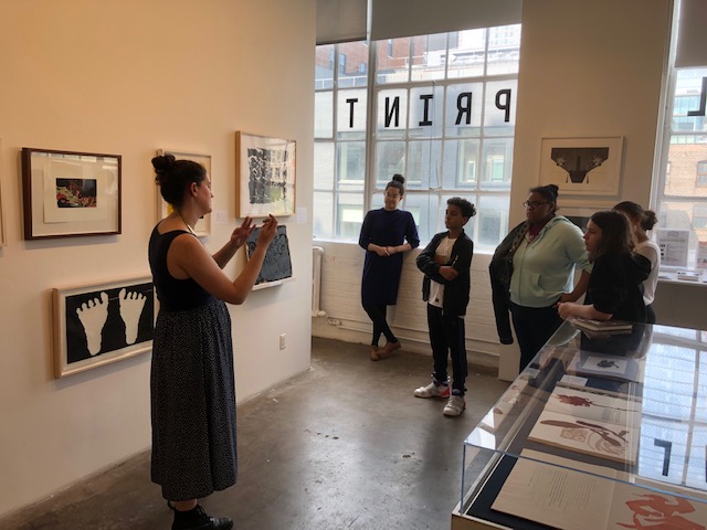Several students listen to a woman speaking, while standing in an exhibition space. Several materials are arranged in a vatrine and several framed artworks hang on the opposite wall. Behind the students are large open windows opening to a cityscape, and one of the windows has the word 