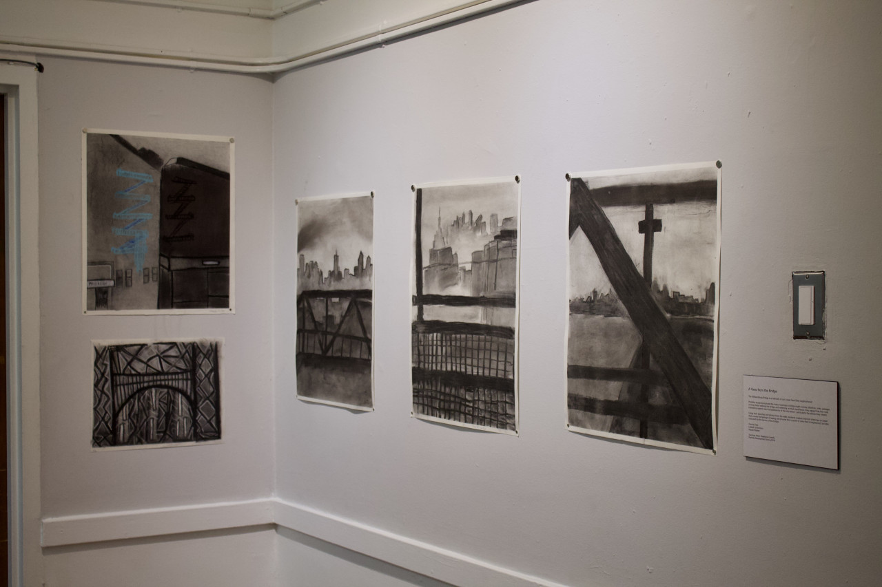 Five rectangular black-and-white drawings of cityscapes appear exhibited on a wall in the corner of a room, with a didactic panel under a lightswitch to their right, titled "A View from the Bridge"