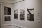Five rectangular black-and-white drawings of cityscapes appear exhibited on a wall in the corner of a room, with a didactic panel under a lightswitch to their right, titled 
