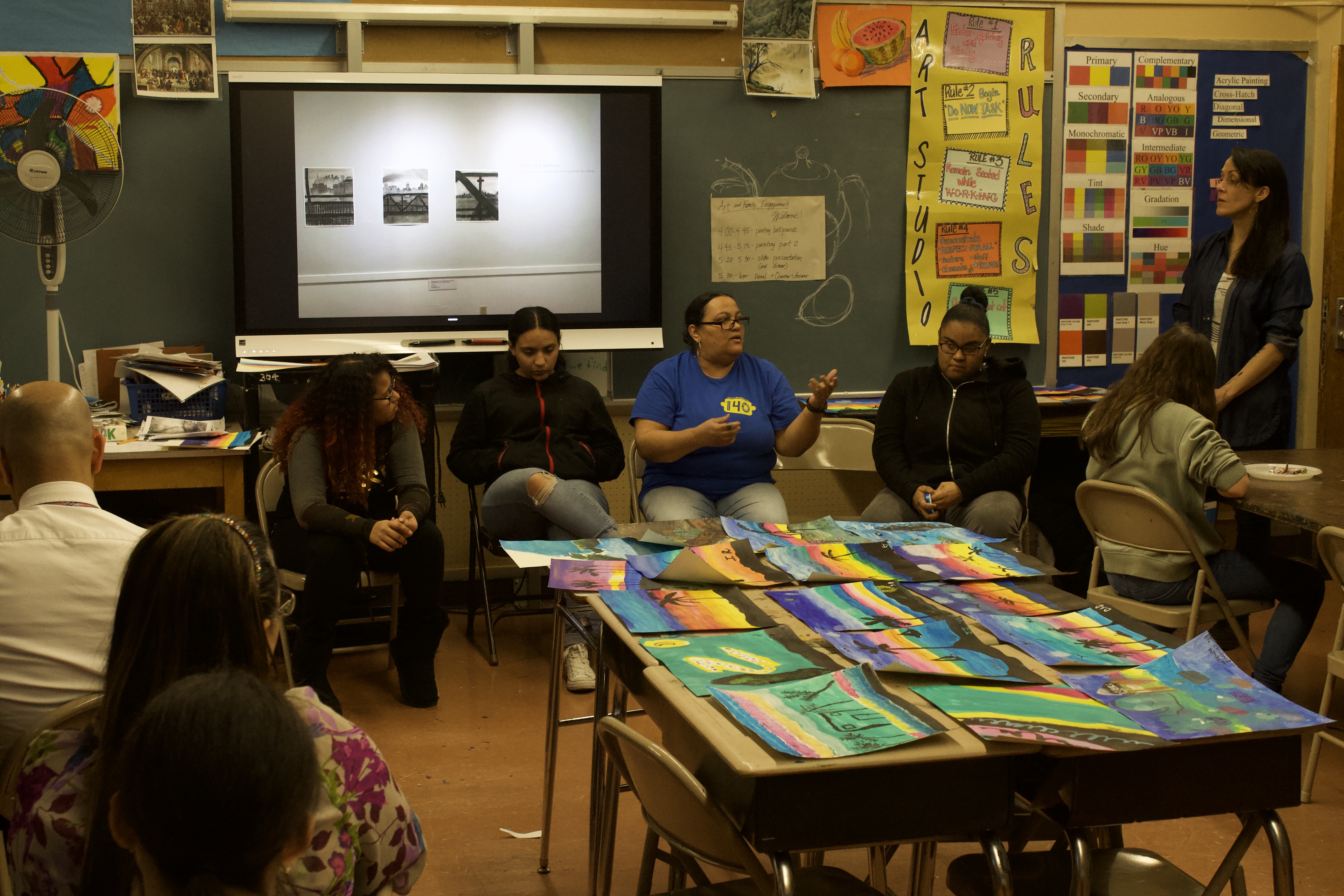 A classroom with several adults sitting around the perimeter. In the center, colorful student artworks are arranged across several desks.