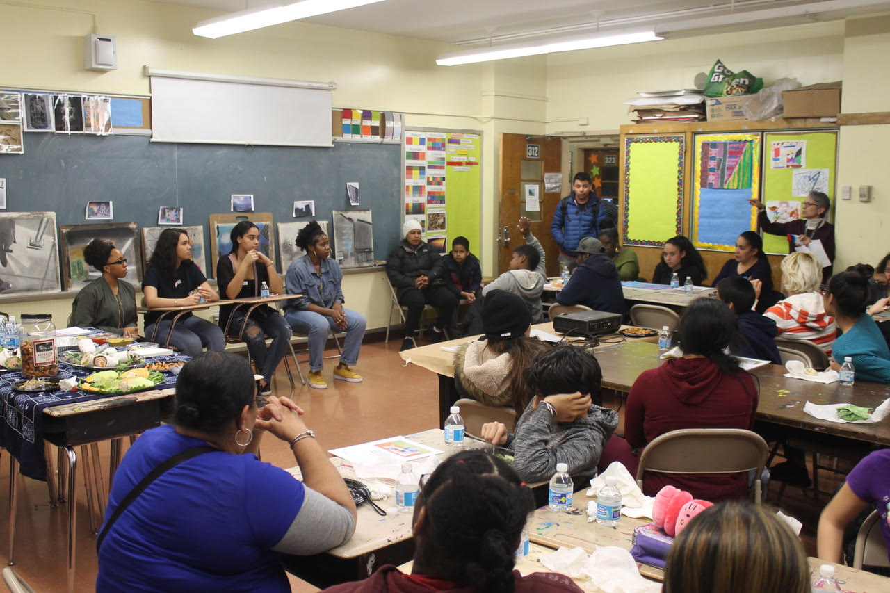 A classroom setting with several students and adults sitting across the room at desks and tables, looking at a teacher speaking to them in a corner of the room