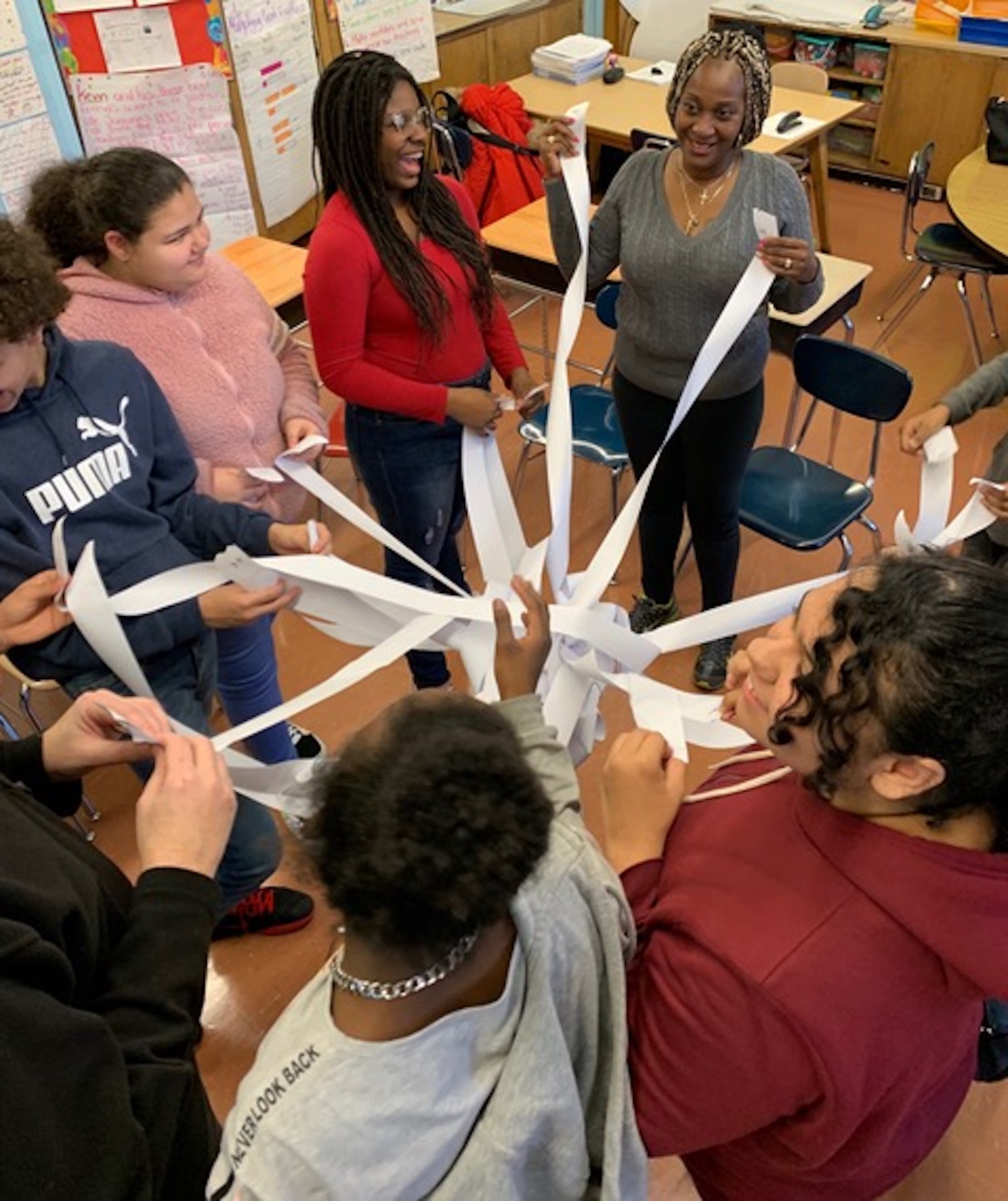 Photograph of students and a teacher in a grey sweater stand in a classroom. They are smiling and holding the ends of white strips of paper which are woven into a tight knot in the center.