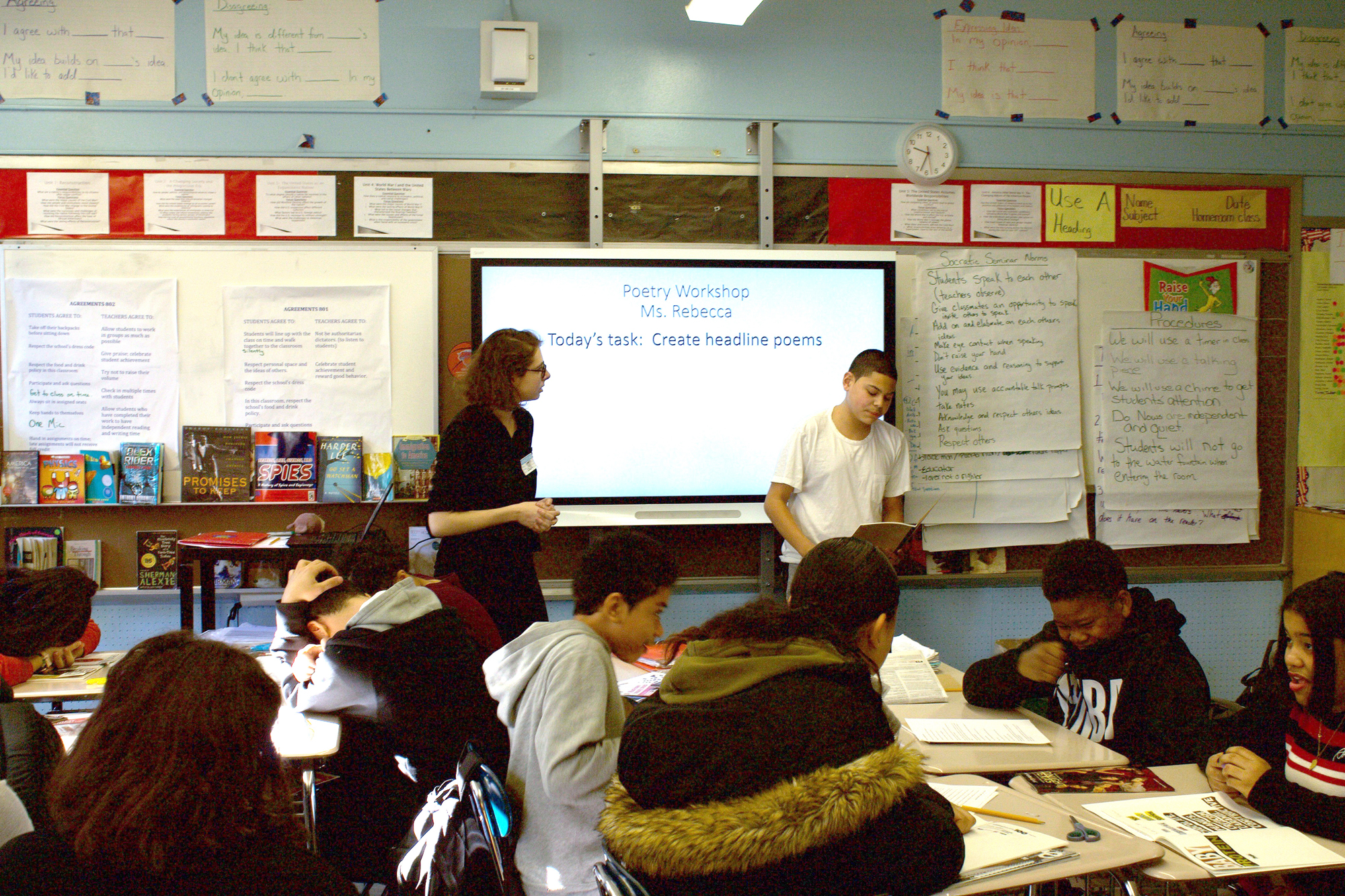 Two people stand at the front of a full classroom in front of a projector screen that says, 
