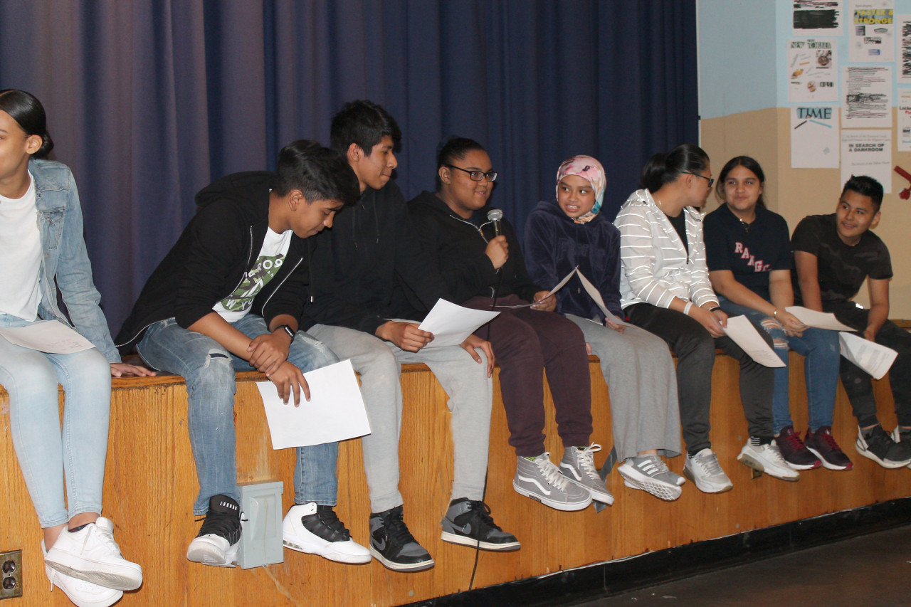 Eight people sit in a row on a wooden stage in front of a navy blue curtain. They are holding pieces of paper, one speaks into a microphone.