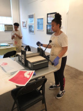 Two people in aprons use a printmaker. One stands in the foreground, using the machine, while the other stands to the left in the background.