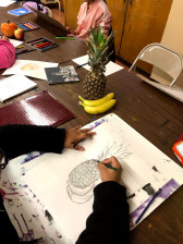 A pineapple and two bananas sit on a table with a person's hands in the midst of drawing the fruit with pencil at the center of the image.