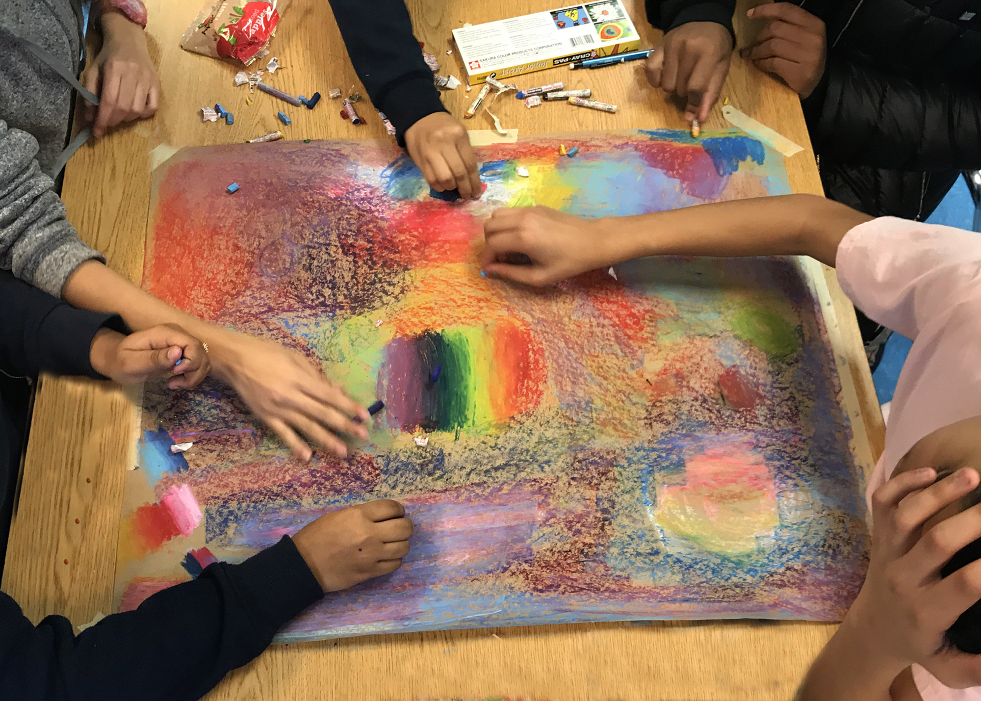 Five people's hands work on a colorful, abstract drawing with the drawing at the center of the image on a wooden table.
