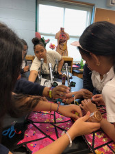 Several students stand around a large desk working on mixed-media artworks. One reaches for something from another student towards the camera.