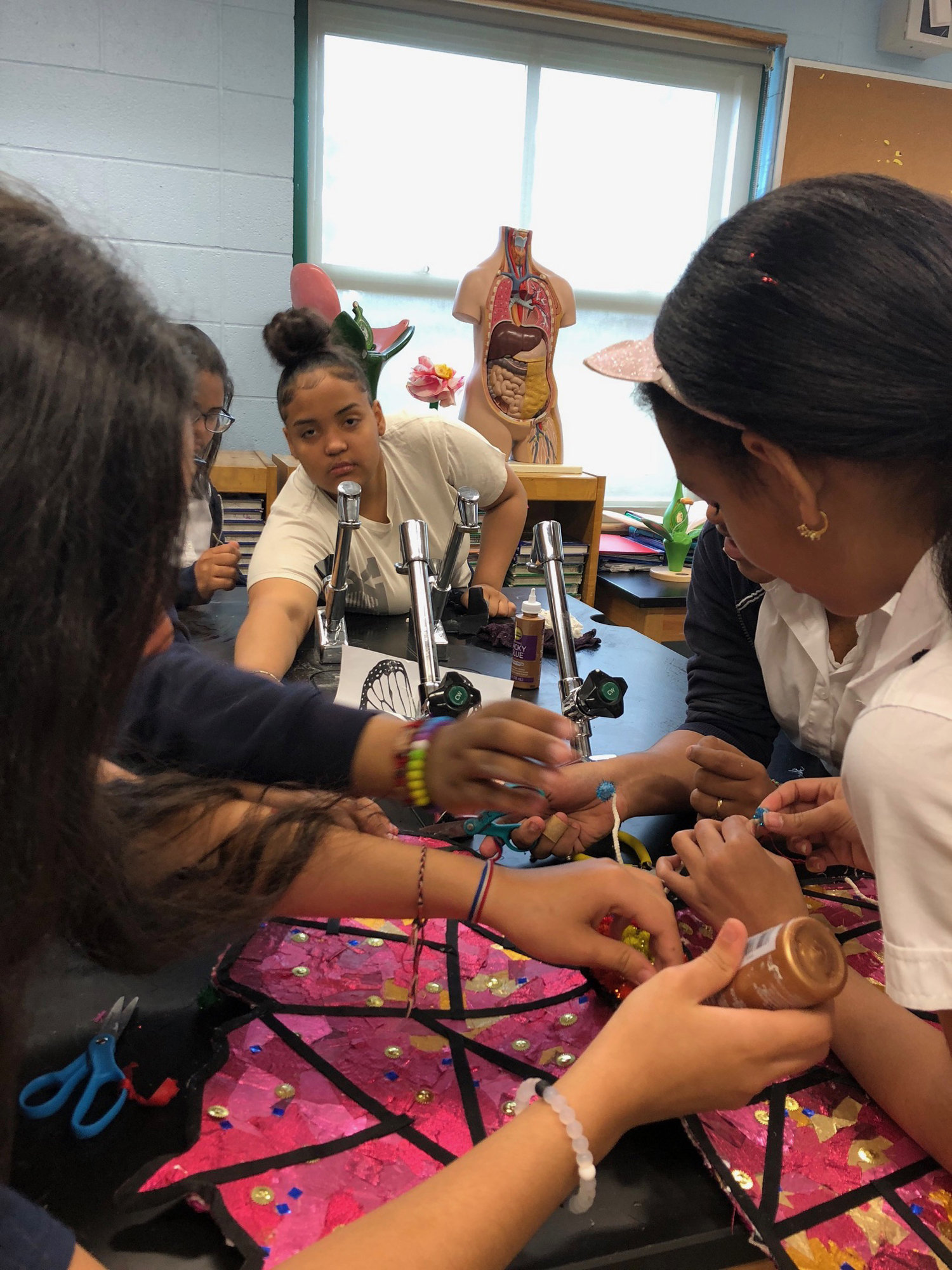 Several students stand around a large desk working on mixed-media artworks. One reaches for something from another student towards the camera.