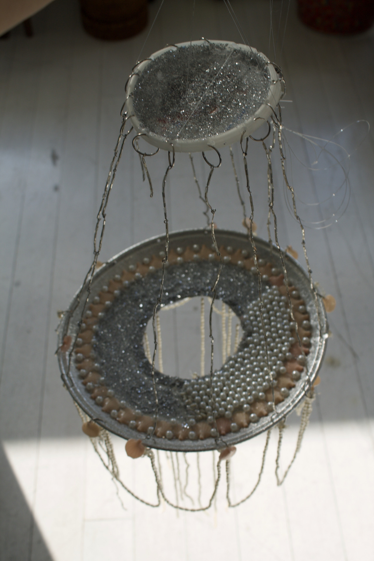 A handmade chandelier, suspended in the air. It has two disks hanging apart, sewn together with silver thread. A number of beads adorn the lower disk and string hanging down below.