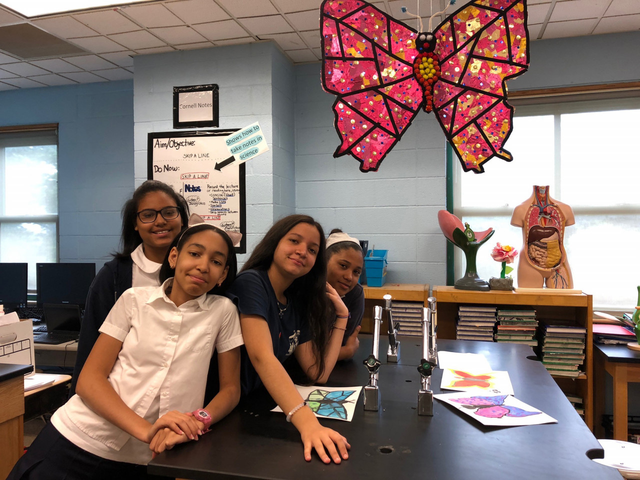 Four people pose for a photo, leaning on a table with three drawings of butterflies splayed across the surface. A papier mache butterfly composed of saturated pink and gold paper and red, yellow, black and white cotton balls hangs above the people at the top right of the image.