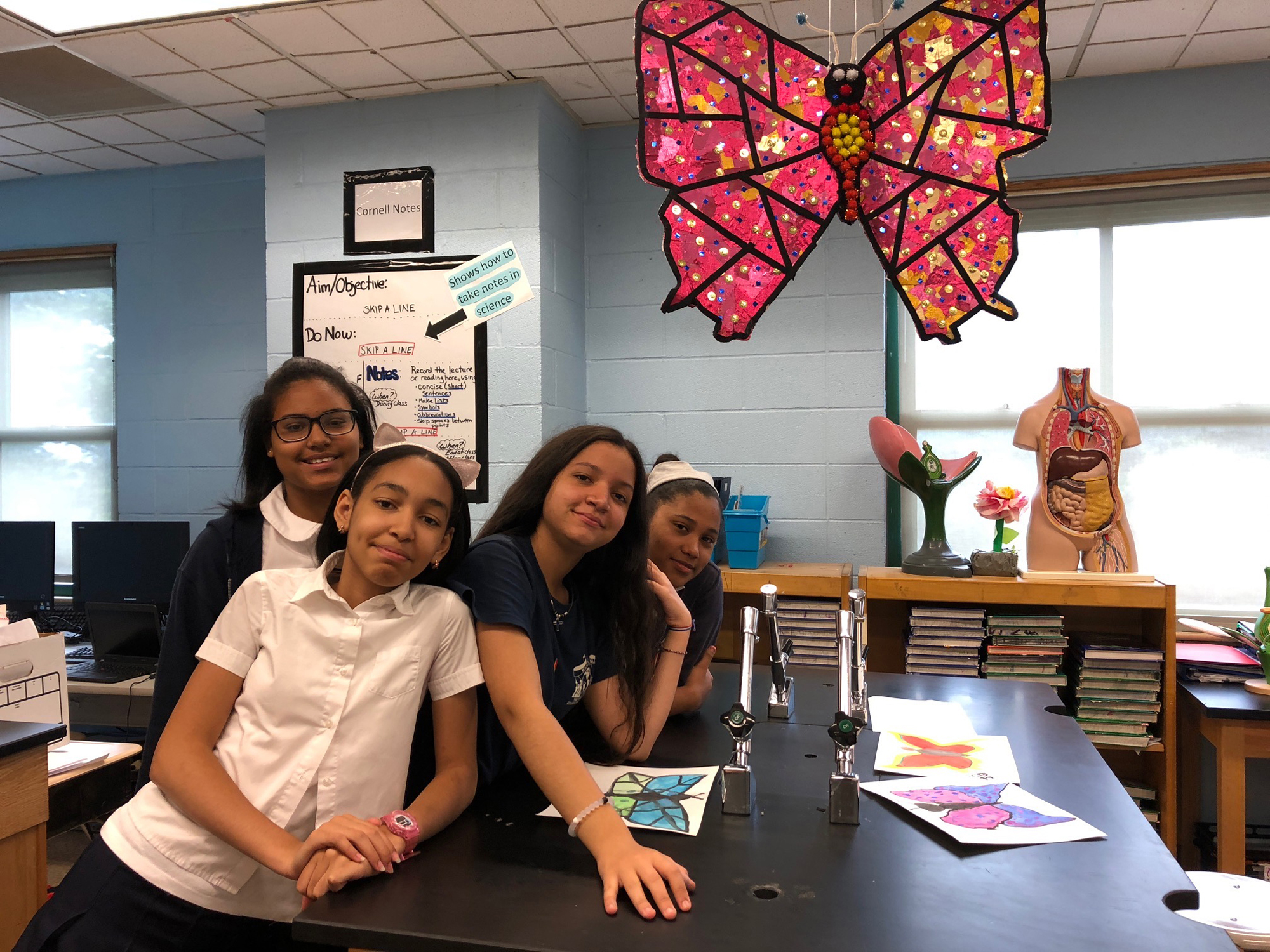Four people pose for a photo, leaning on a table with three drawings of butterflies splayed across the surface. A papier mache butterfly composed of saturated pink and gold paper and red, yellow, black and white cotton balls hangs above the people at the top right of the image.