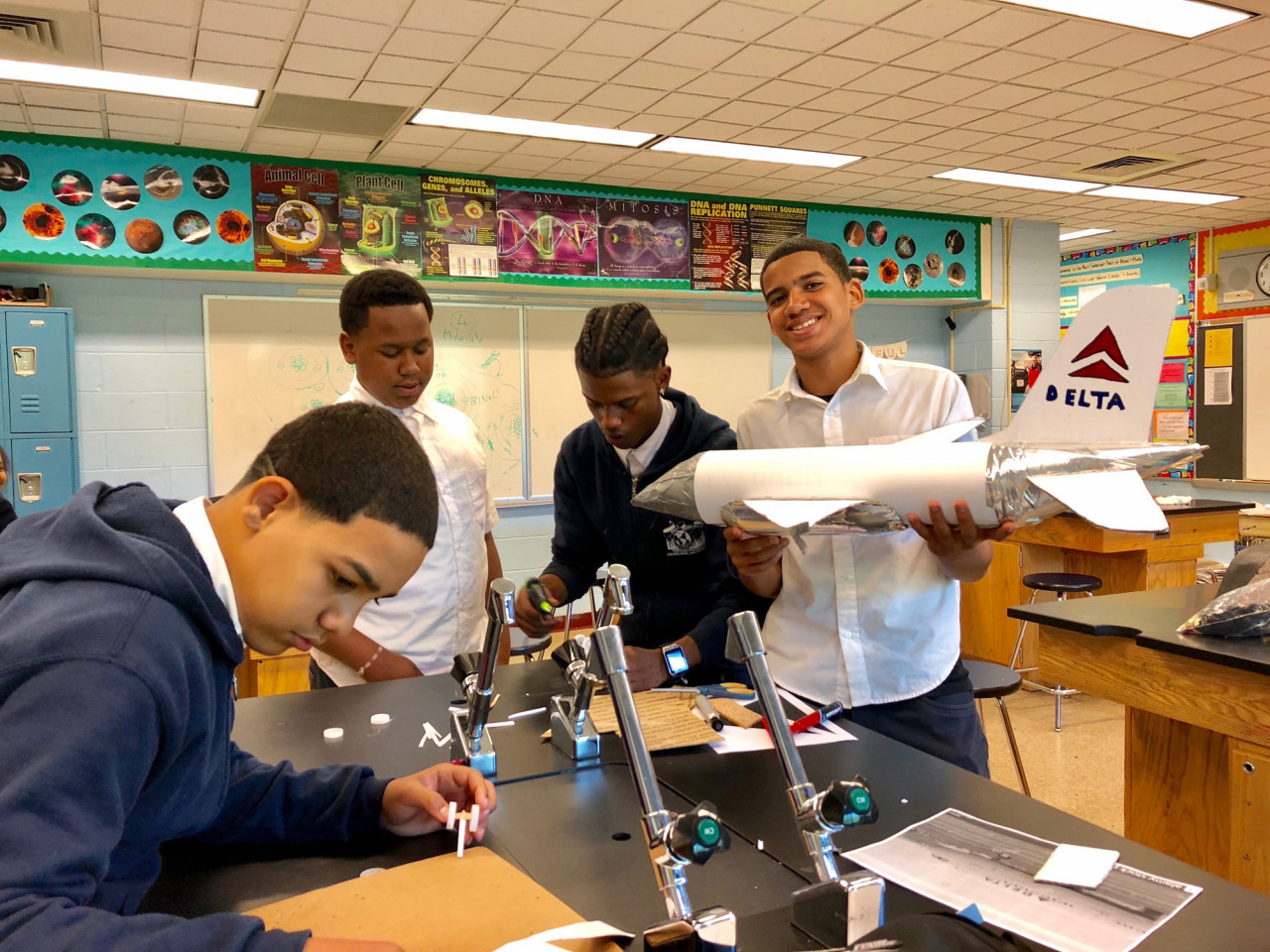 Several students stand around a large science room desk. One holds a model of a Delta airplane, smiling for the camera. The rest are busy at work.