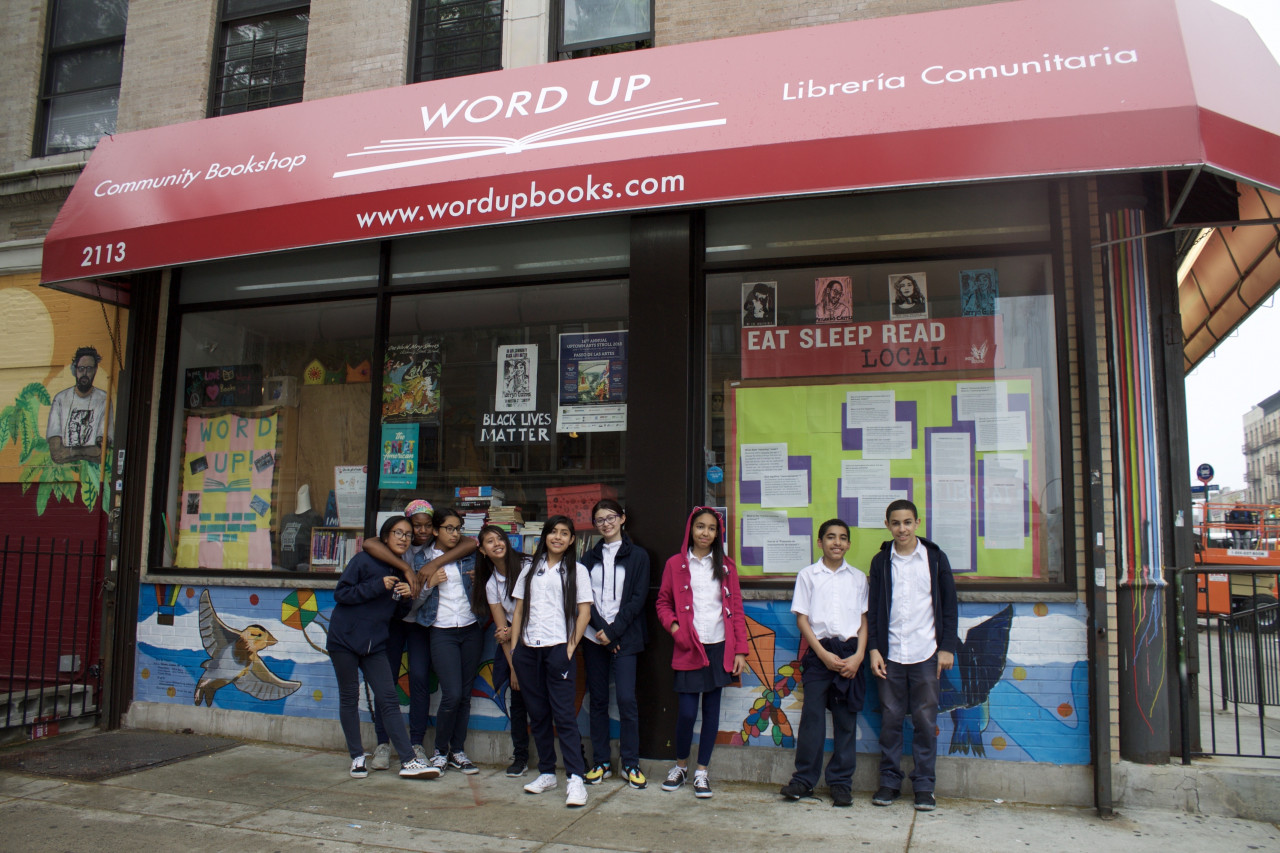 Students stand outside of a storefront in New York - Word Up Community Bookshop. In the window are several signs with Black Lives Matter and "Eat Sleep Read Local" slogans
