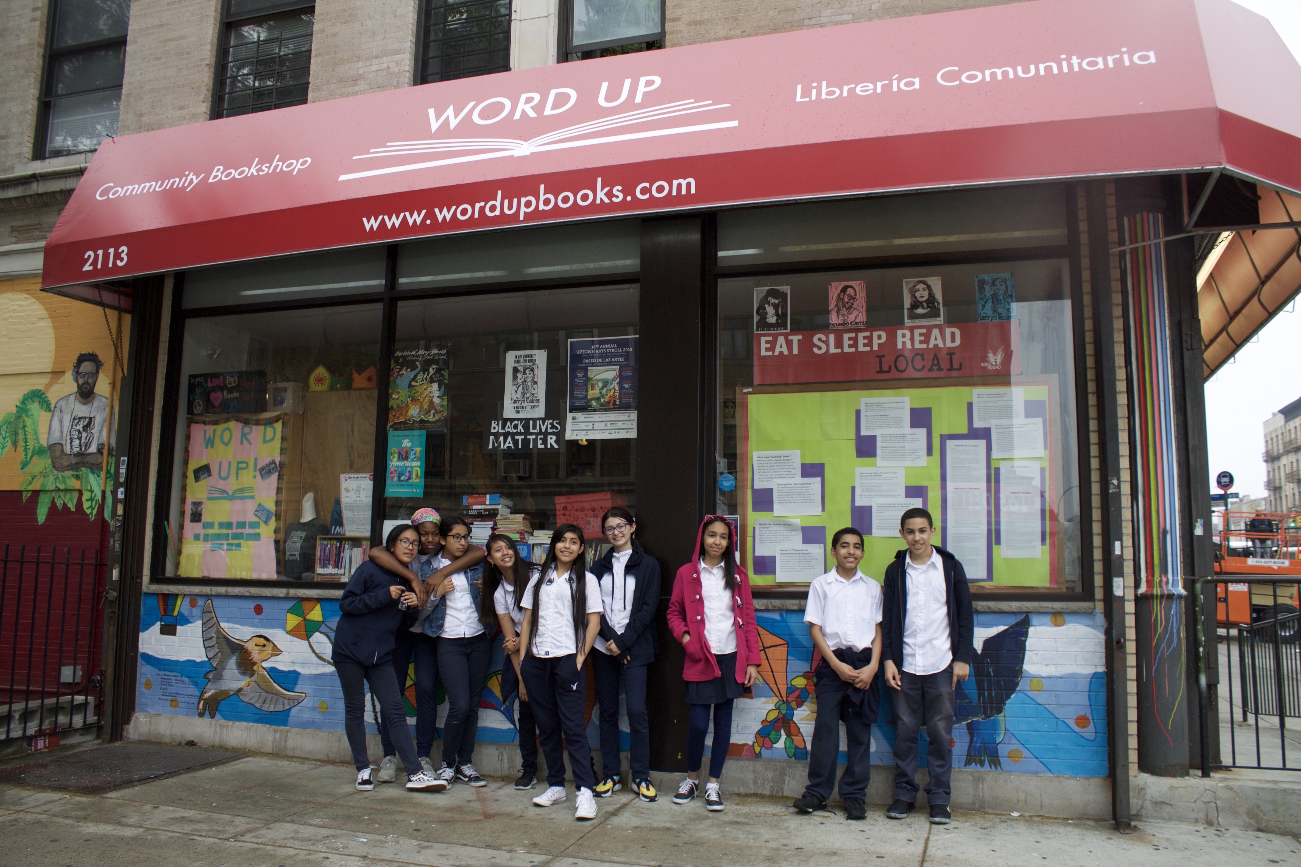 Students stand outside of a storefront in New York - Word Up Community Bookshop. In the window are several signs with Black Lives Matter and 