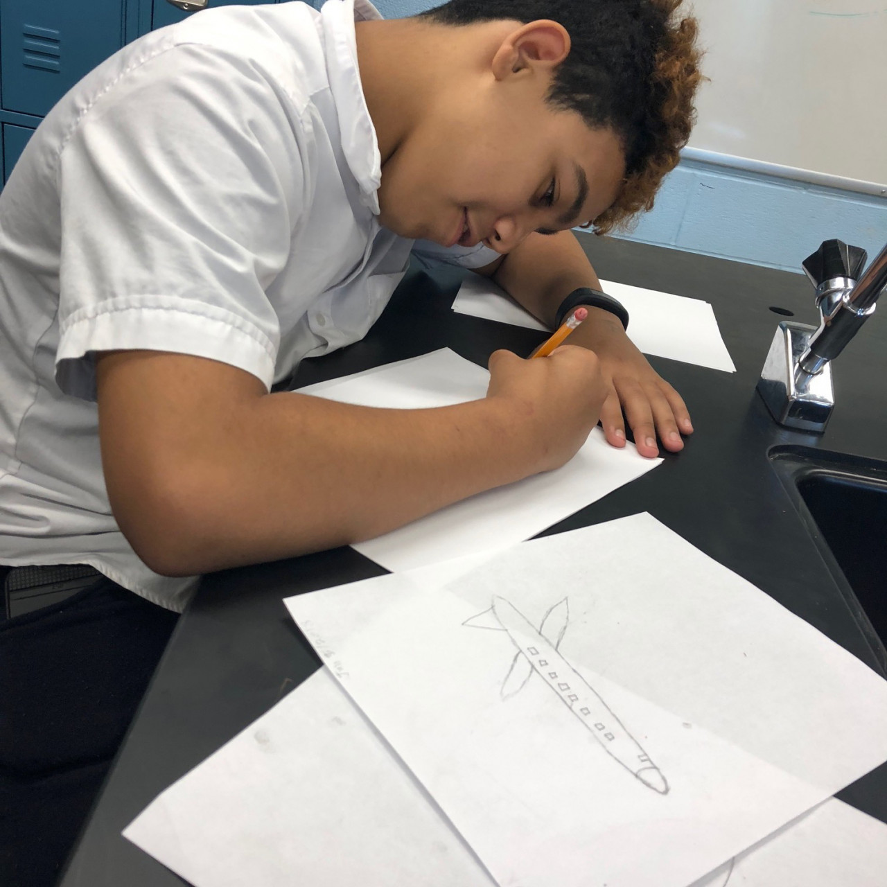 A student sitting at a science room desk with a sink, drawing with a pencil on several sheets of paper. One sheet, foregrounded, has an outline of an airplane