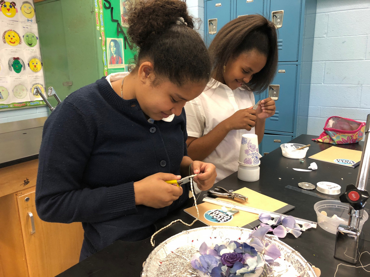 Two girls stand, cutting threads, while working on a handmade chandelier. The chandelier has a small number of flower petals, with a purple rose in the center