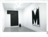 Two paintings and a small indistinguishable sculpture are visible on adjacent walls with wall text on a wall to the left of them in a white-walled gallery space. The painting on the right shows a darkened American flag next with a block of black extending from the lefthand corner and the small sculpture to its right on the bottom. The painting on the left reads 