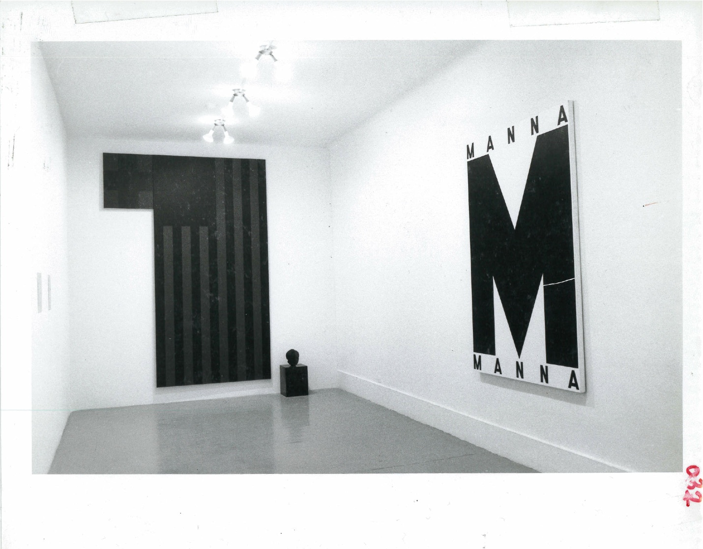 Two paintings and a small indistinguishable sculpture are visible on adjacent walls with wall text on a wall to the left of them in a white-walled gallery space. The painting on the right shows a darkened American flag next with a block of black extending from the lefthand corner and the small sculpture to its right on the bottom. The painting on the left reads 