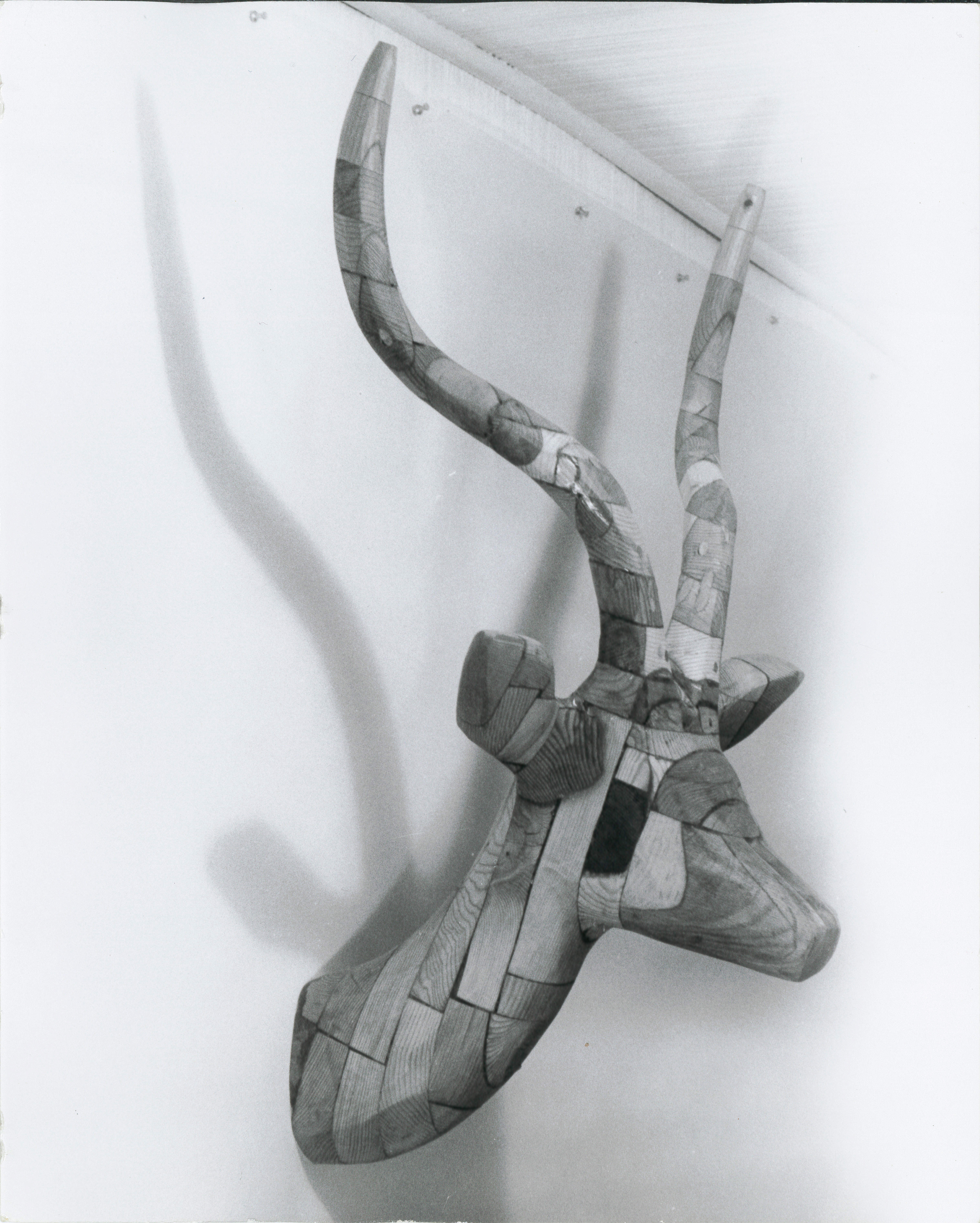 A black and white photograph of a wooden bust of a horned deer like animal, attached to a white wall.