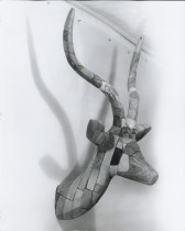 A black and white photograph of a wooden bust of a horned deer like animal, attached to a white wall.
