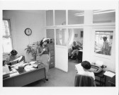 Black and white photo of various figures in an office space. The set and costumes are in the style of the 1950s. One woman sits at a desk looking through papers, one woman sits at a typewriter. Three other figures are also in the space in casual poses.