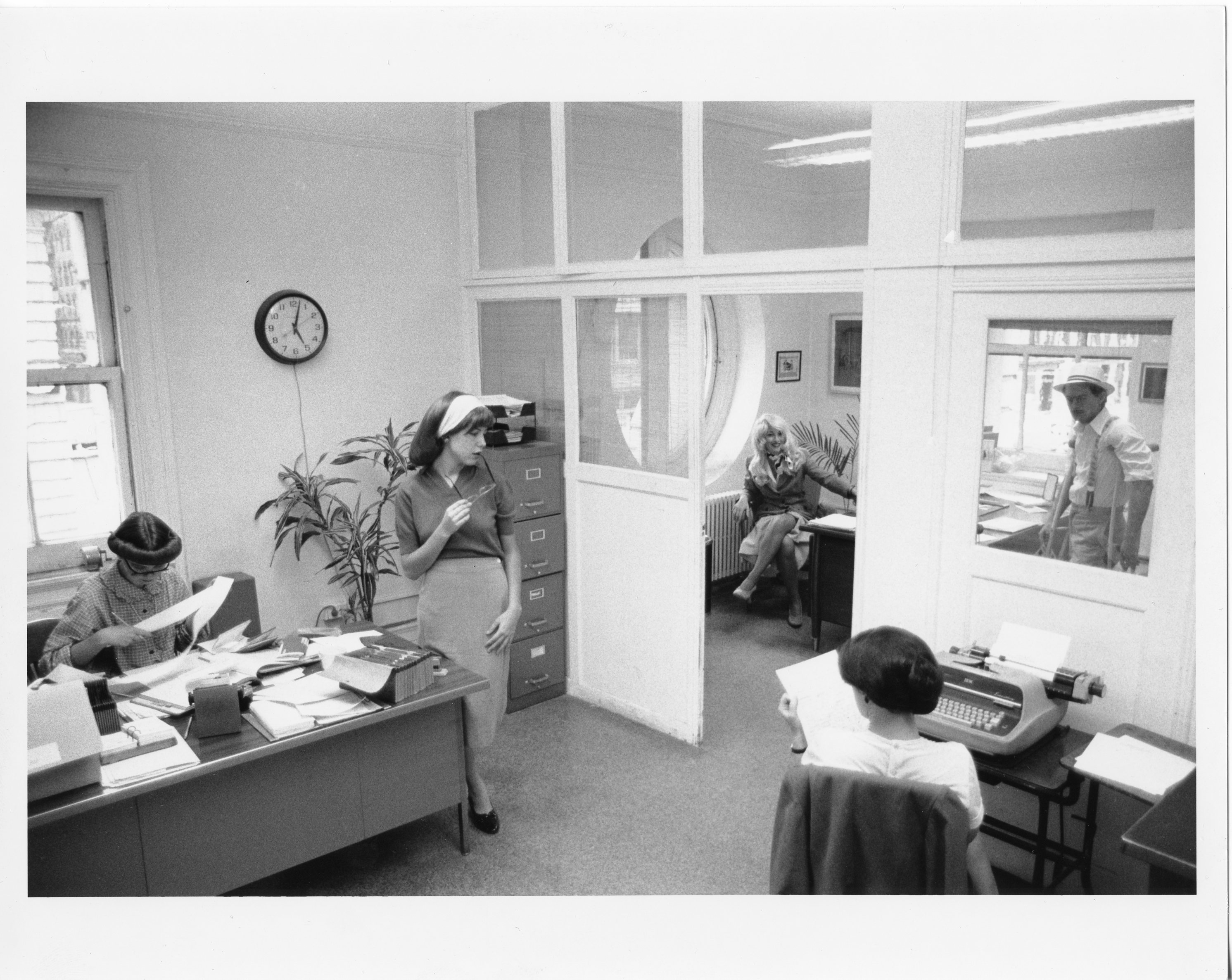 Black and white photo of various figures in an office space. The set and costumes are in the style of the 1950s. One woman sits at a desk looking through papers, one woman sits at a typewriter. Three other figures are also in the space in casual poses.