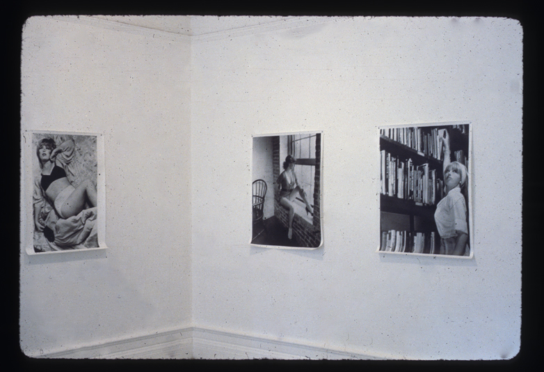 Three black and white photographs hang on white walls in the corner of a room. The photos depict a woman in various positions: laying down, reaching up to a bookshelf, and sitting on a ledge gazing out a window.
