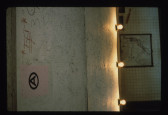 A white wall with the Artists Space logo affixed. Light bulbs are affixed to the edge of the wall. In the background a piece hangs on the wall.