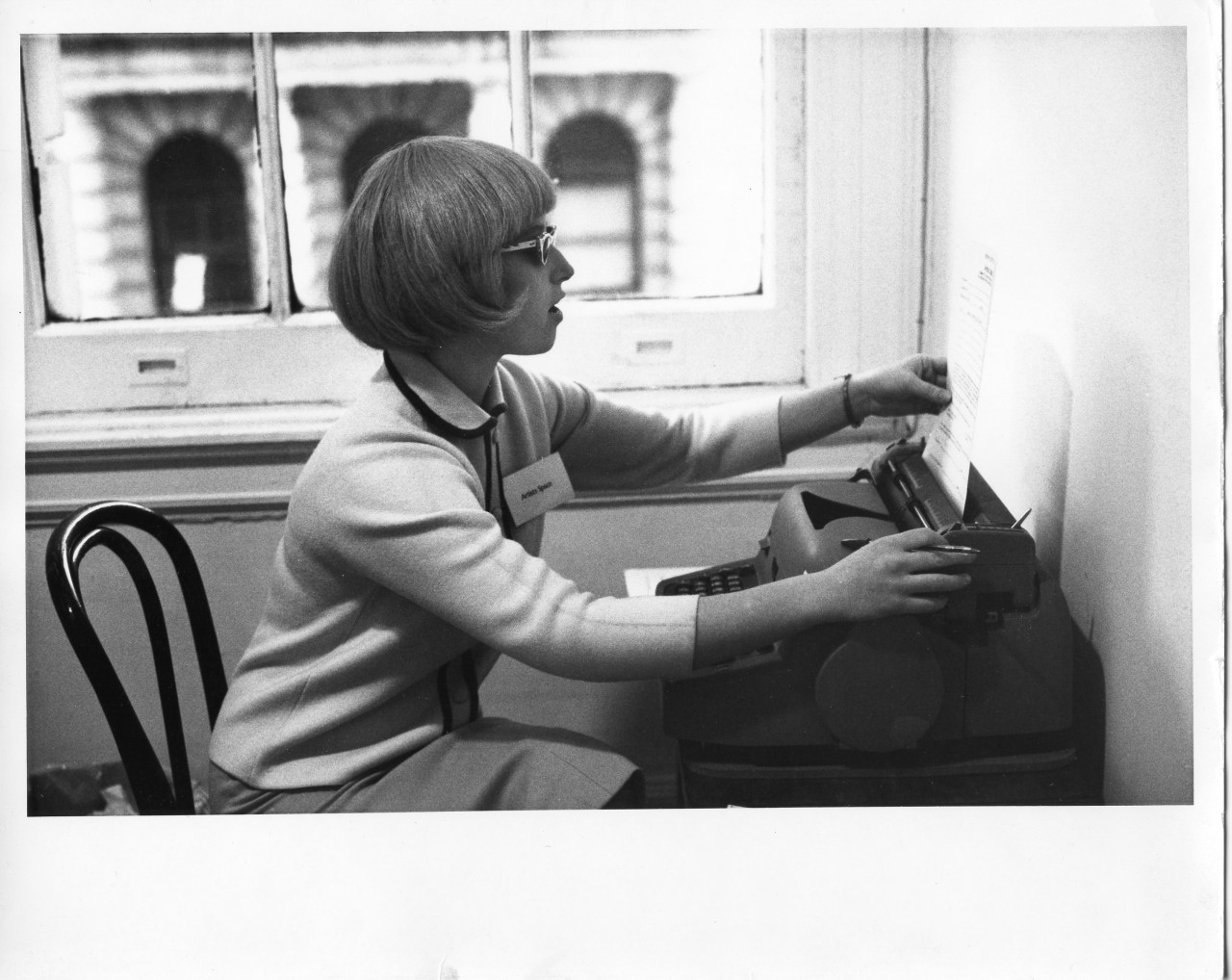 A woman sits in front of a typewriter inserting a piece of paper. She is dressed in 1950s fashion.