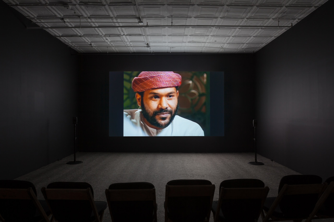 A dimly lit, empty room filled with low chairs. A projection illuminates a dark wall opposite the chairs and displays an image of a man with tan skin and facial hair, wearing a keffiyeh.