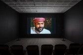 A dimly lit, empty room filled with low chairs. A projection illuminates a dark wall opposite the chairs and displays an image of a man with tan skin and facial hair, wearing a keffiyeh.