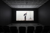 A photograph of an empty room, dimly lit with chairs in the background, where a projection is playing on the wall. The projection displays an image of a man holding up the Iraqi flag.