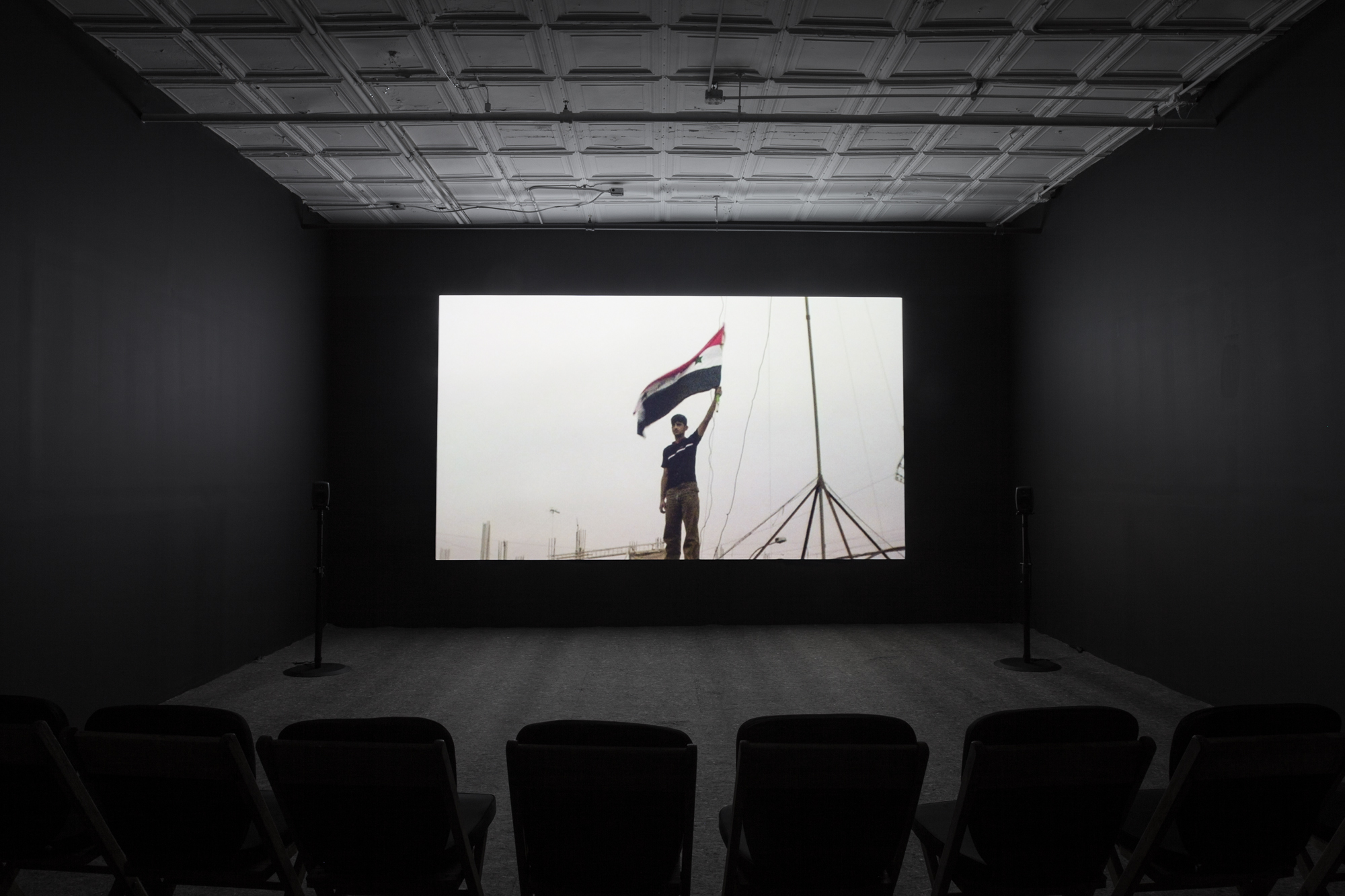 A photograph of an empty room, dimly lit with chairs in the background, where a projection is playing on the wall. The projection displays an image of a man holding up the Iraqi flag.