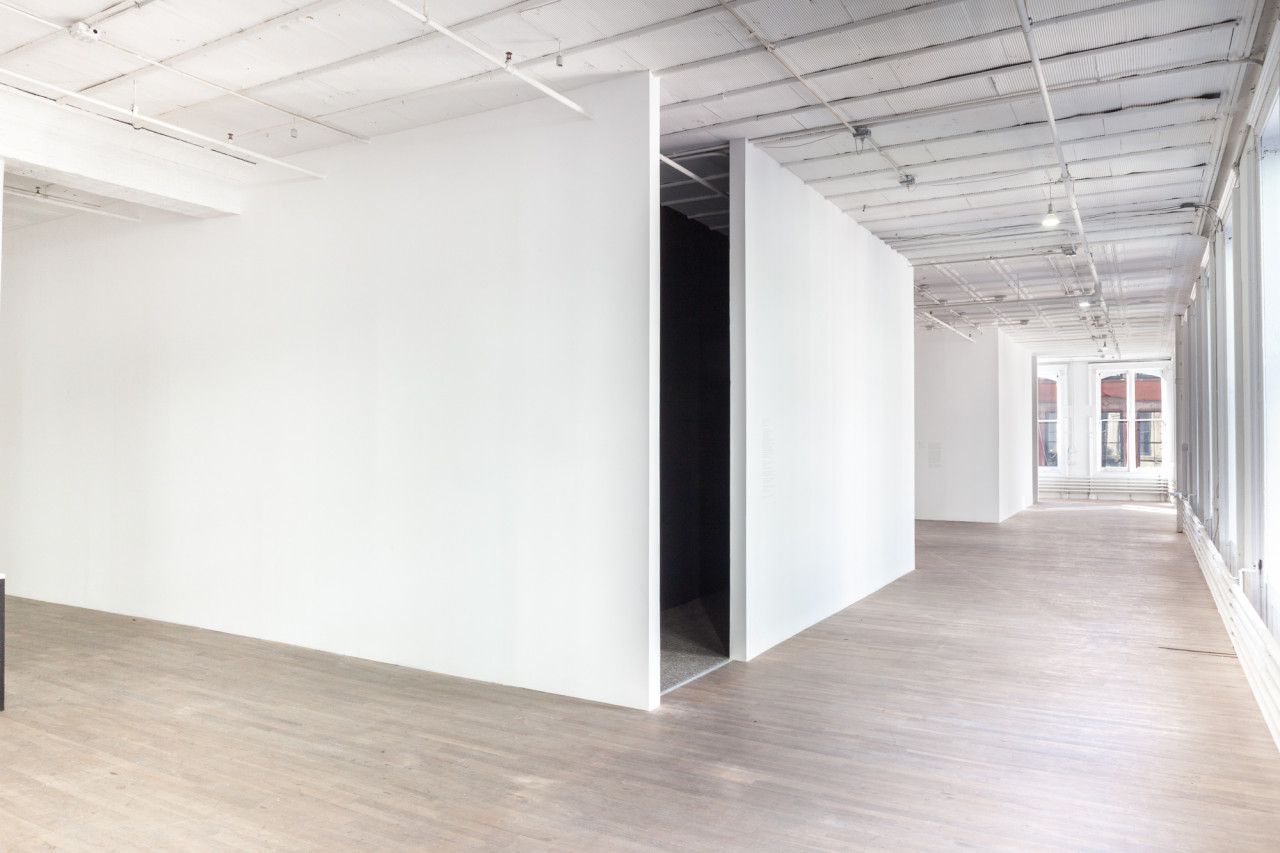 Several blank, white walls in a large gallery space with wooden flooring. An opening emerges at the corner of two intersecting walls at the image