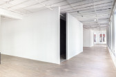 Several blank, white walls in a large gallery space with wooden flooring. An opening emerges at the corner of two intersecting walls at the image's center, leading into a dark space. Rows of windows line the right wall of the gallery.