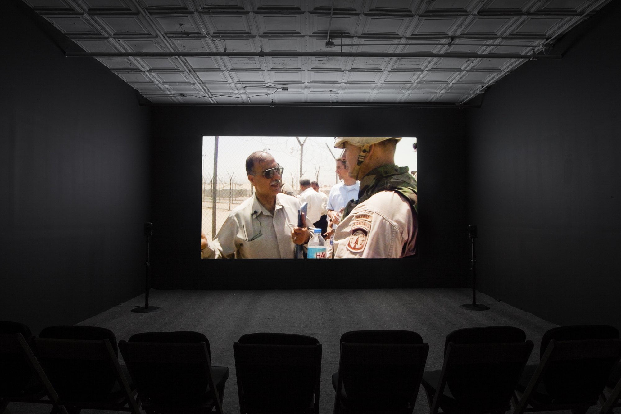 A photograph of an empty room, dimly lit with chairs in the background, where a projection is playing on the wall. The projection displays an image of a man with tan skin and facial hair, wearing aviators, talking to a soldier.