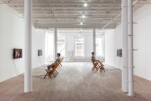 Two figures sit in wooden chairs in a light-filled gallery space, wearing headphones and facing television monitors installed on opposites sides of the room.