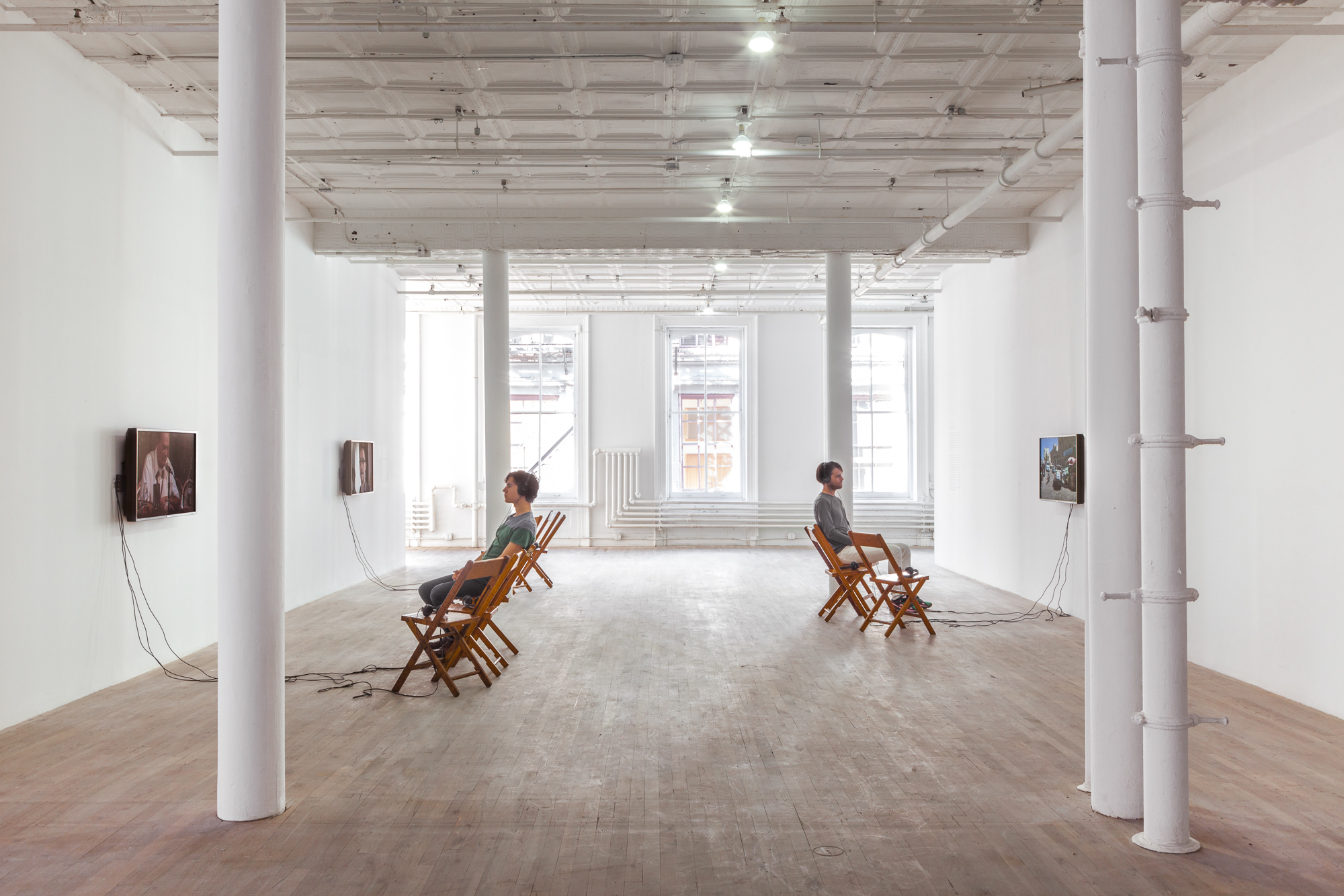 Two figures sit in wooden chairs in a light-filled gallery space, wearing headphones and facing television monitors installed on opposites sides of the room.