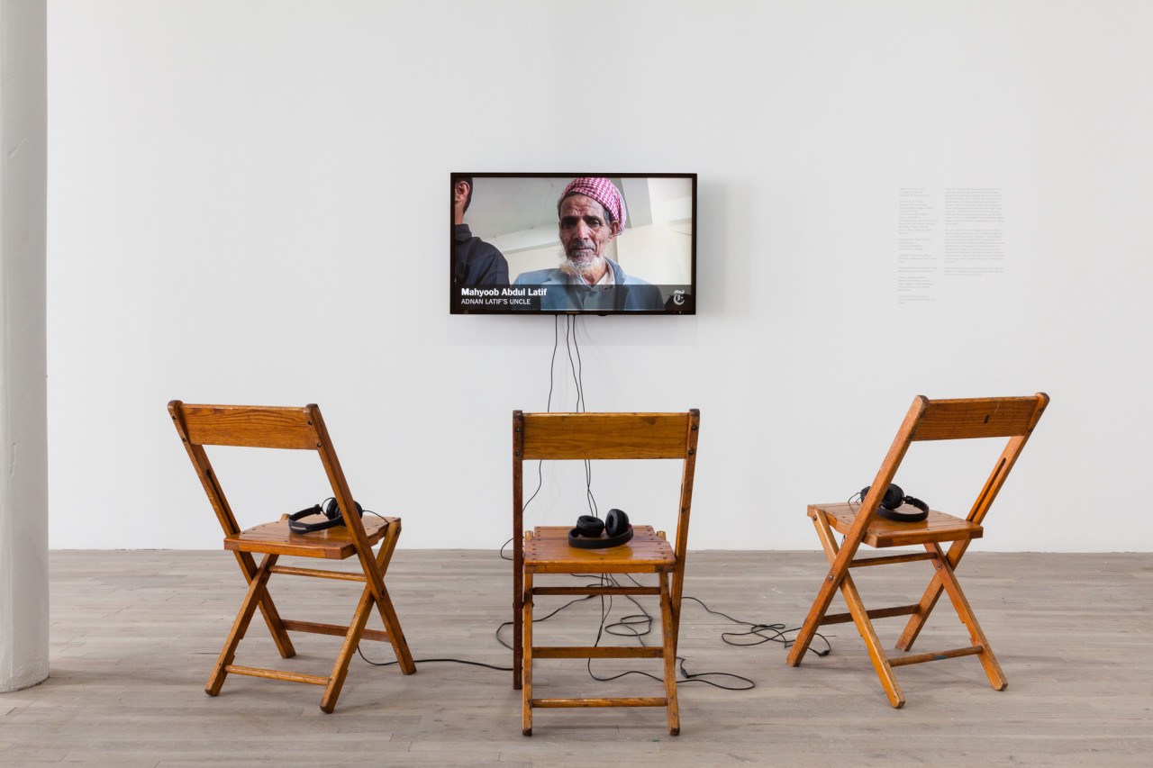 Three wooden chairs with headphones on top of them face a television monitor, which displays a figure wearing a keffiyeh. White text underneath the figure