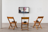 Three wooden chairs with headphones on top of them face a television monitor, which displays a figure wearing a keffiyeh. White text underneath the figure's face reads: 