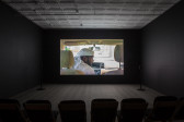 A dimly lit, empty room filled with low chairs. A projection illuminates a dark wall opposite the chairs. The projection shows the interior of a vehicle, where a man wearing a keffiyeh sits in the front seat of the vehicle and leans over to the passenger seat.