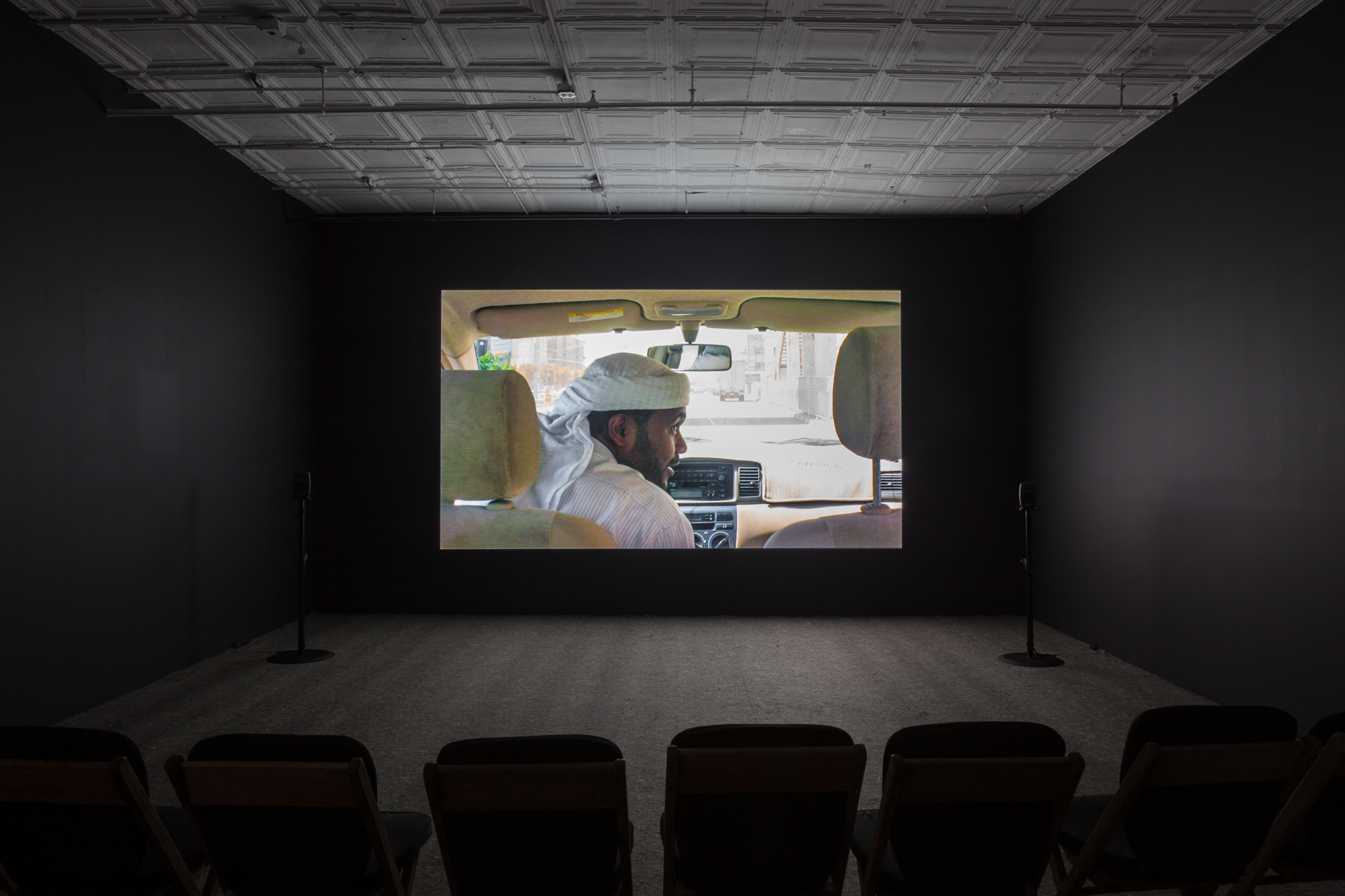A dimly lit, empty room filled with low chairs. A projection illuminates a dark wall opposite the chairs. The projection shows the interior of a vehicle, where a man wearing a keffiyeh sits in the front seat of the vehicle and leans over to the passenger seat.