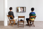 Two individuals seated in two wooden chairs while a third in between them holds a single headphone. They are seated in front of a TV monitor displaying Edward Snowden's image.