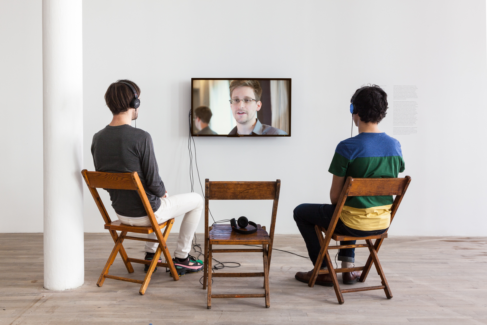 Two individuals seated in two wooden chairs while a third in between them holds a single headphone. They are seated in front of a TV monitor displaying Edward Snowden's image.