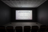 A photograph of an empty room, dimly lit with chairs in the background, where a projection is playing on the wall. The projection displays an image of the Hamdan vs. Rumsfield Supreme Court Case.