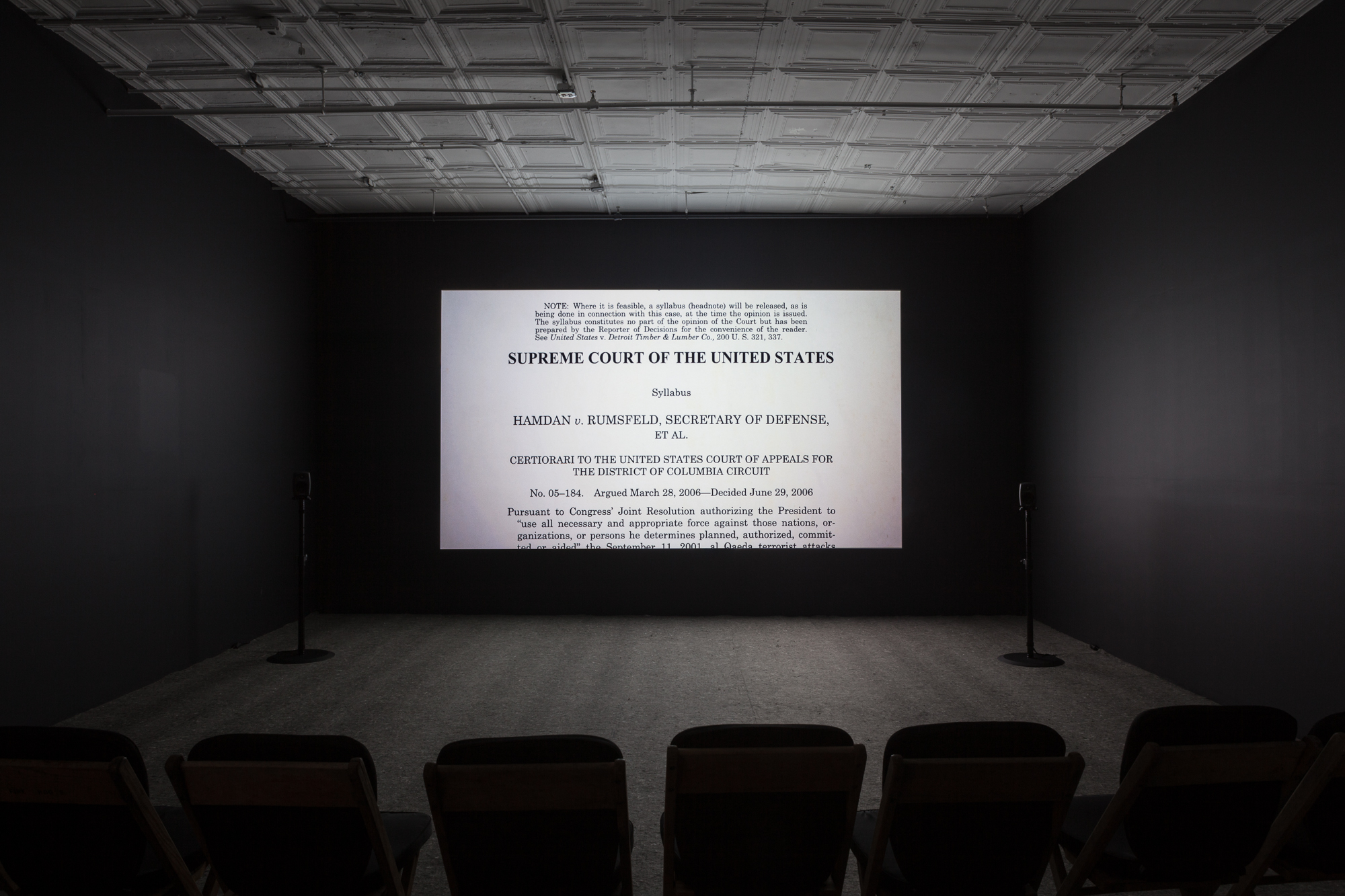 A photograph of an empty room, dimly lit with chairs in the background, where a projection is playing on the wall. The projection displays an image of the Hamdan vs. Rumsfield Supreme Court Case.
