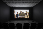 A photograph of an empty room, dimly lit with chairs in the background, where a projection is playing on the wall. The projection displays an image a soldier interacting with civilians. The caption reads, 
